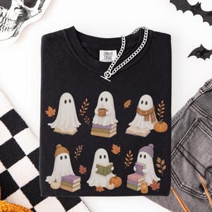 Black shirt featuring six cute bookish ghosts reading books with pumpkins, stacked books, fall leaves, scarves, hats, and a cozy drink.