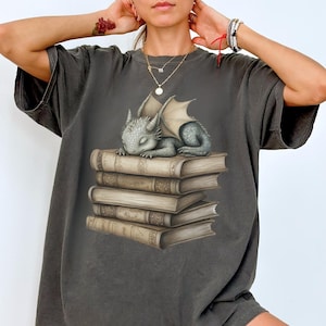 May include: An oversized t-shirt in a dark grey colour featuring a graphic of a small grey dragon sleeping on top of a stack of old books.