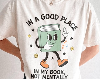 May include: Cream-colored t-shirt with the text "IN A GOOD PLACE" above a cartoon book character. The book has a face, arms, and legs, and the text "IN MY BOOK, NOT MENTALLY" is below. The design includes stars, musical notes, and flowers.