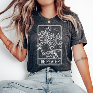 May include: Dark gray t-shirt featuring a white graphic of an open book with roses and the text "THE READER." The design is framed by a rectangular border with the Roman numeral XII at the top. The shirt is made of a soft material.