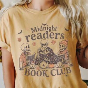 May include: A mustard yellow t-shirt featuring a graphic of three skeletons reading books at a table with tea, a teapot, a pumpkin, and candles. The text reads Midnight readers BOOK CLUB, surrounded by small bats, ghosts, and autumn leaves.