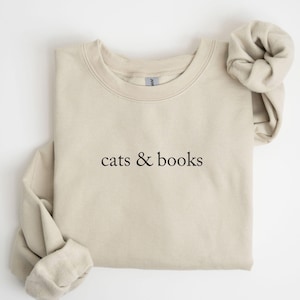 Puede incluir: Sudadera beige con la frase "cats & books" en negro. La camiseta de manga larga está doblada cuidadosamente, con una manga enrollada. La tela parece suave y cómoda, adecuada para uso casual.