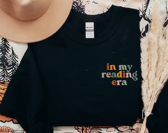 May include: Black crewneck sweatshirt with the text "in my reading era" in colorful lettering. A tan hat and dried ornamental grass are also in the image. The sweatshirt is a Gildan brand.