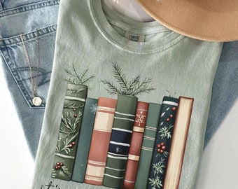 May include: Sage green t-shirt featuring a graphic of illustrated books and the text "it's reading season." The books are in shades of green, red, and blue, with decorative elements. A gold necklace, light blue jeans, and a tan hat are also in the image.