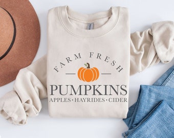 Puede incluir: Una sudadera beige con el texto "FARM FRESH PUMPKINS APPLES - HAYRIDES - CIDER" y un pequeño gráfico de calabaza naranja.