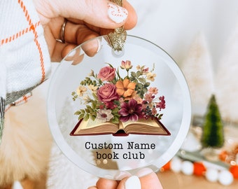 May include: A clear, round Christmas ornament with a floral design and an open book illustration. The ornament features a bouquet of pink, orange, and burgundy flowers. The text "Custom Name book club" is printed below the book.