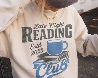 May include: A brown baseball cap with "La Petite Paris" embroidered on the front. A cream sweatshirt with "Late Night Reading Club Estd 2025" and a blue coffee cup graphic. The person is holding a coffee drink.