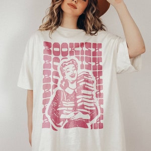 Könnte beinhalten: Ein weißes Oversize-T-Shirt mit einer pinkfarbenen Grafik einer Frau, die einen Stapel Bücher hält. Die Grafik enthält das Wort "BOOKISH" in pinkfarbenen Buchstaben.