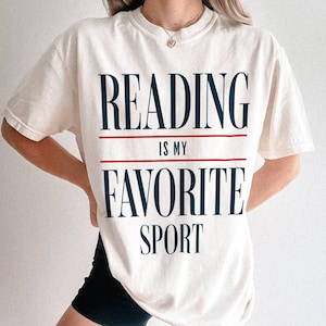 May include: A white t-shirt with the text "READING IS MY FAVORITE SPORT" printed in blue and red.