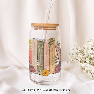 May include: A clear glass tumbler with a bamboo lid and a straw. The tumbler is decorated with a design of books and flowers. The book titles include "Killers of the Flower Moon", "The Great Gatsby", "To Kill a Mockingbird", and "The Grapes of Wrath".