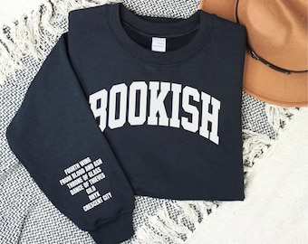 May include: A navy blue sweatshirt with the word "BOOKISH" in large white block letters across the chest. The left sleeve features a list of book titles in white text. A brown hat rests nearby on a patterned surface.