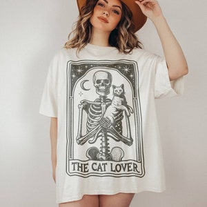 May include: Cream-colored t-shirt featuring a black and white graphic of a skeleton holding a cat, with the text "THE CAT LOVER" below. The design includes a crescent moon and stars. The shirt has short sleeves and a relaxed fit.