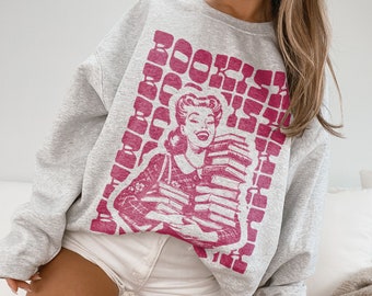 Puede incluir: Una sudadera gris con un gráfico rosa de una mujer sosteniendo una pila de libros. El gráfico incluye el texto "BOOKISH".