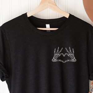 May include: Black t-shirt with a white skeleton hand heart graphic. The shirt is hanging on a wooden hanger. A tan hat and a beige purse with flowers are also visible.