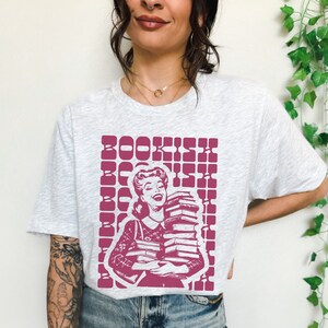 May include: A white t-shirt with a pink graphic of a woman holding a stack of books. The graphic has the word "BOOKISH" repeated in a grid pattern.