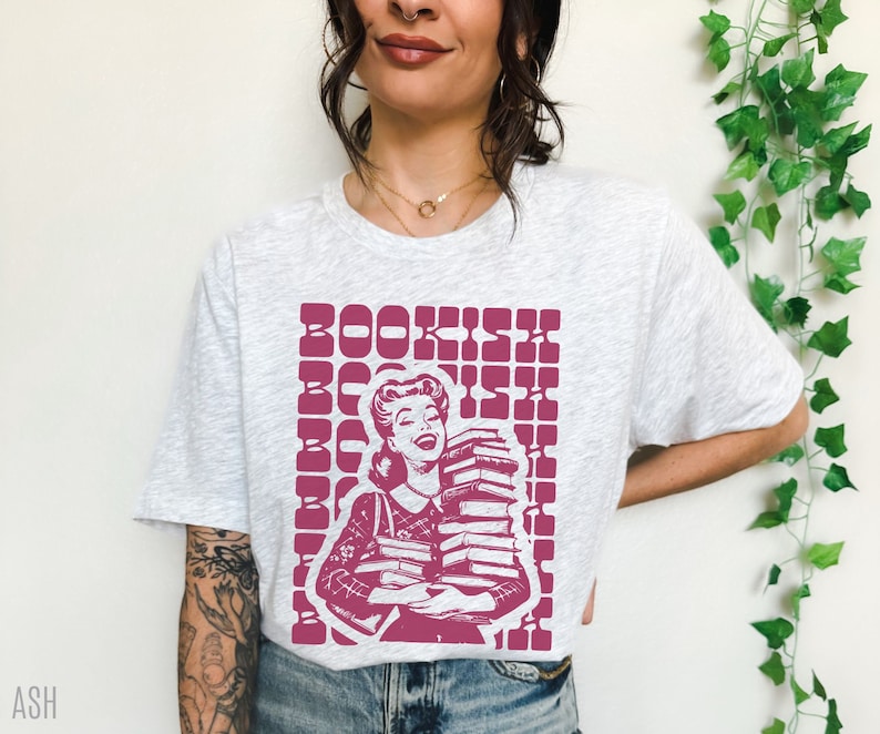 May include: A white t-shirt with a pink graphic of a woman holding a stack of books. The graphic has the word "BOOKISH" repeated in a grid pattern.
