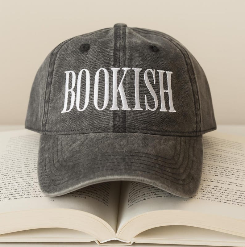 May include: A gray baseball cap with the word "BOOKISH" embroidered in white. The cap is sitting on top of an open book. The hat has a curved brim and an adjustable strap.