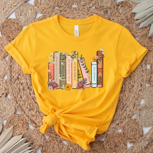 May include: A yellow t-shirt with a graphic of a stack of books with floral accents. The book titles include "The Fault in Our Stars", "ACOTAR", "From Blood and Ash", "It Ends With Us", and "Crescent City".