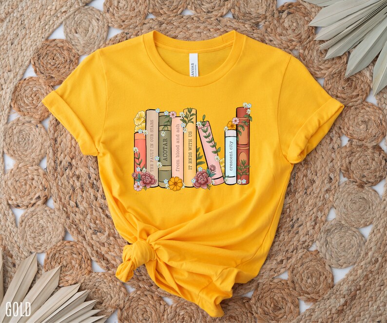 May include: A yellow t-shirt with a graphic of a stack of books with floral accents. The book titles include "The Fault in Our Stars", "ACOTAR", "From Blood and Ash", "It Ends With Us", and "Crescent City".