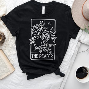 May include: Black t-shirt featuring a white graphic of a tarot card design. The design includes an open book with roses and the text "THE READER." The top is styled with a knot at the waist.