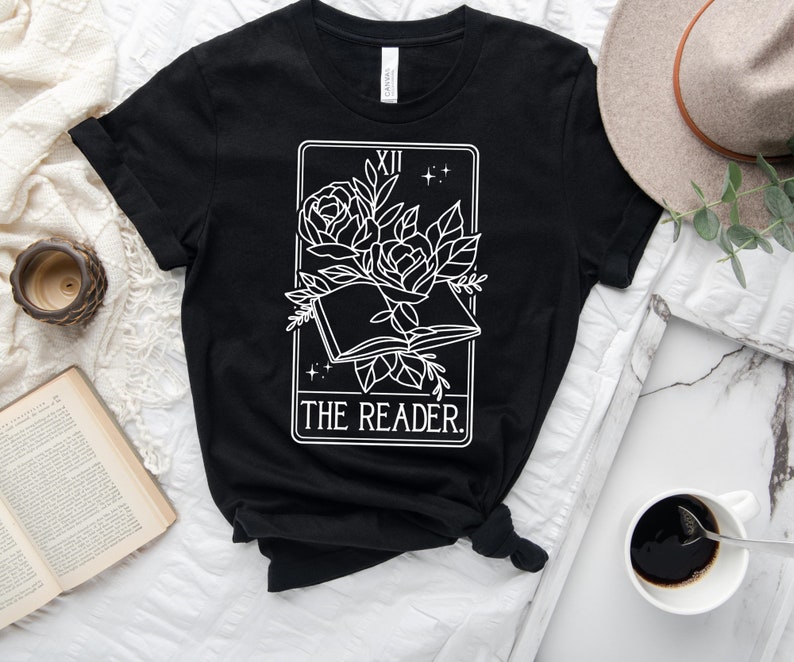 May include: Black t-shirt featuring a white graphic of a tarot card design. The design includes an open book with roses and the text "THE READER." The top is styled with a knot at the waist.
