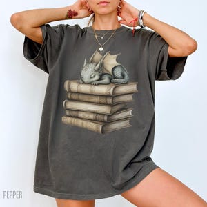 May include: Dark grey t-shirt featuring a sleeping baby dragon on a stack of books. The dragon is grey with folded wings. The books are tan with detailed illustrations. The word "PEPPER" is printed at the bottom left.