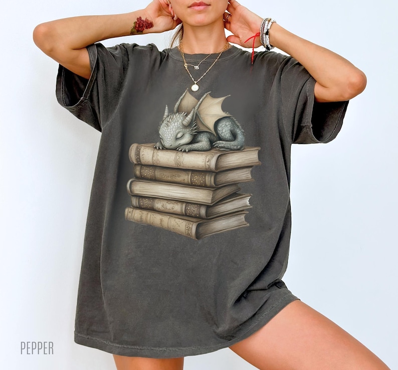 May include: Dark grey t-shirt featuring a sleeping baby dragon on a stack of books. The dragon is grey with folded wings. The books are tan with detailed illustrations. The word "PEPPER" is printed at the bottom left.