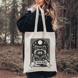 May include: Cream-colored tote bag featuring a black and white illustration of a raccoon reading a book under a night sky with stars and a moon. The text "THE TRASH READER" is displayed below the raccoon.