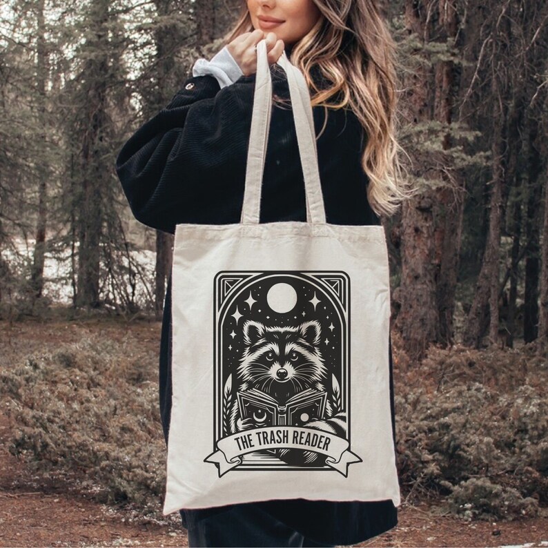 May include: Cream-colored tote bag featuring a black and white illustration of a raccoon reading a book under a night sky with stars and a moon. The text "THE TRASH READER" is displayed below the raccoon.