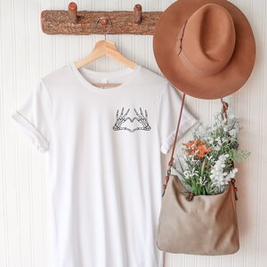 May include: A white t-shirt with a black skeleton hand heart design. The shirt is hanging on a wooden hanger. A brown hat and a tan purse filled with flowers are also visible.
