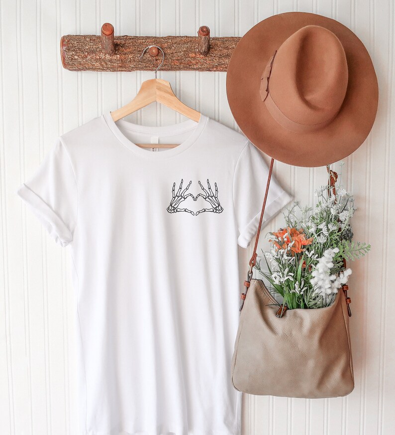 May include: A white t-shirt with a black skeleton hand heart design. The shirt is hanging on a wooden hanger. A brown hat and a tan purse filled with flowers are also visible.