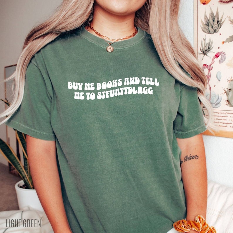 May include: Light green t-shirt with the text "BUY ME BOOKS AND TELL ME TO STFUATTDLAGG" in white, a round gold necklace, and a gold bracelet. The shirt is a solid color and has short sleeves.