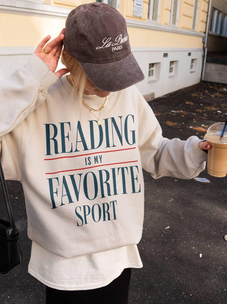 May include: A beige sweatshirt with the text "READING IS MY FAVORITE SPORT" in navy blue and red. The person is wearing a brown baseball cap with "La Poste Paris" embroidered on it. The person is also wearing a gold necklace.