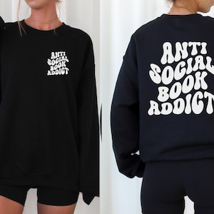 May include: Black sweatshirt with the text "ANTI SOCIAL BOOK ADDICT" in white, on the front left chest and back. The sweatshirt is a long-sleeved crew neck style. The text has a retro, wavy font.