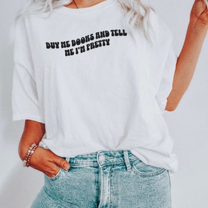 May include: White t-shirt with the text "BUY ME BOOKS AND TELL ME I'M PRETTY" in black, retro-style lettering. The shirt is worn by a person with blonde hair and blue jeans.