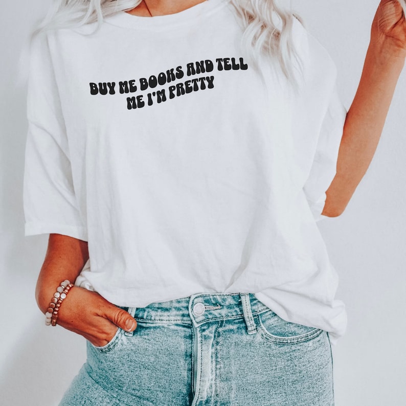 May include: White t-shirt with the text "BUY ME BOOKS AND TELL ME I'M PRETTY" in black, retro-style lettering. The shirt is worn by a person with blonde hair and blue jeans.