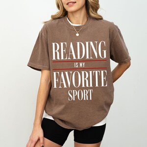 May include: A brown t-shirt with the text "Reading is my favorite sport" printed in white. The text is separated by two red lines.