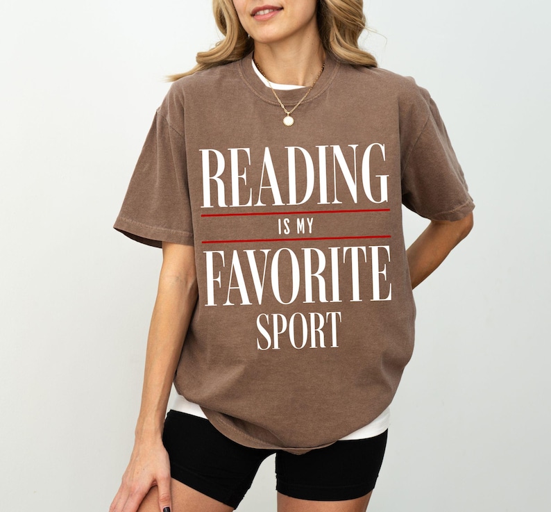 May include: A brown t-shirt with the text "Reading is my favorite sport" printed in white. The text is separated by two red lines.
