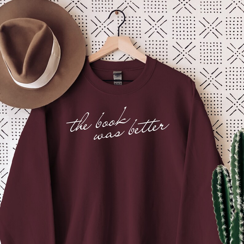 Puede incluir: Una sudadera de cuello redondo color granate con el texto blanco en cursiva "the book was better" impreso en el pecho. La prenda se muestra en una percha de madera, acompa&ntilde;ada de un sombrero marr&oacute;n de ala ancha con banda blanca y un cactus verde.