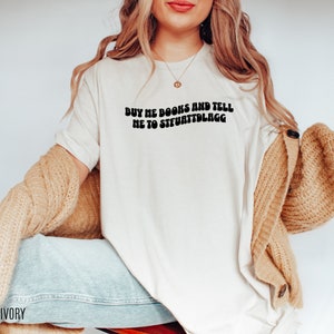 May include: Ivory-colored t-shirt with the text "BUY ME BOOKS AND TELL ME TO STFUATTDLAGG" in black, retro-style font. The shirt is a casual, everyday wear item. The image shows the shirt being worn with a beige cardigan and blue jeans.