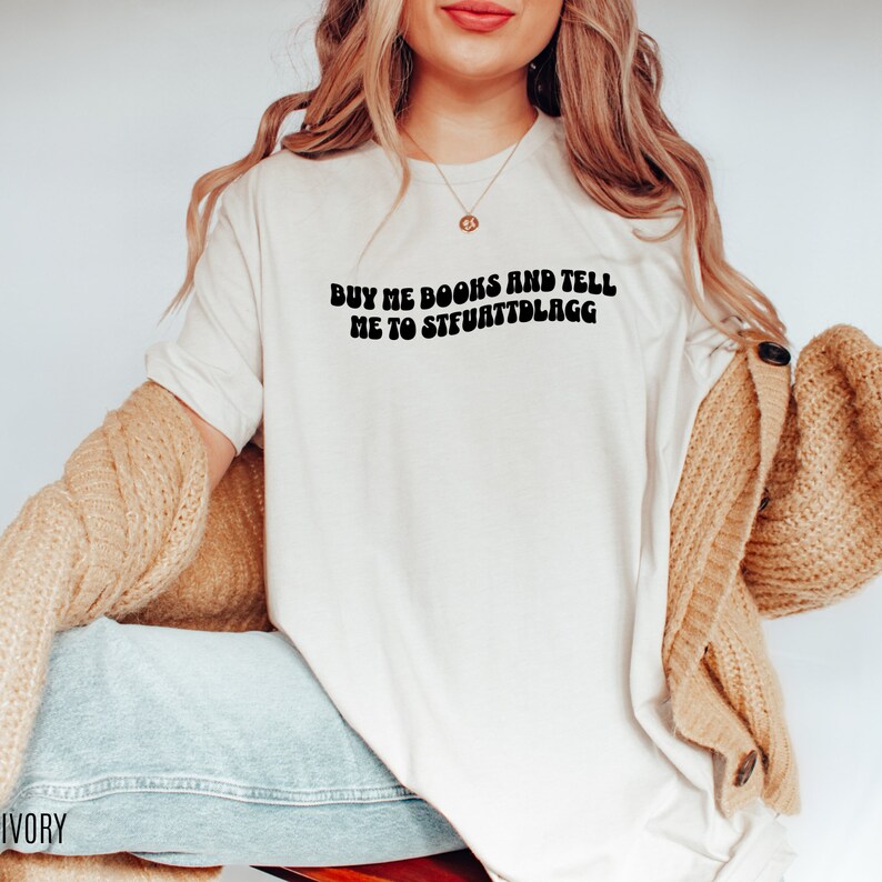 May include: Ivory-colored t-shirt with the text "BUY ME BOOKS AND TELL ME TO STFUATTDLAGG" in black, retro-style font. The shirt is a casual, everyday wear item. The image shows the shirt being worn with a beige cardigan and blue jeans.