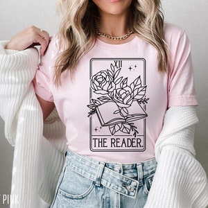 May include: Light pink t-shirt featuring a black and white tarot card design. The design includes an open book, roses, and the text "THE READER." The shirt is made of soft material.
