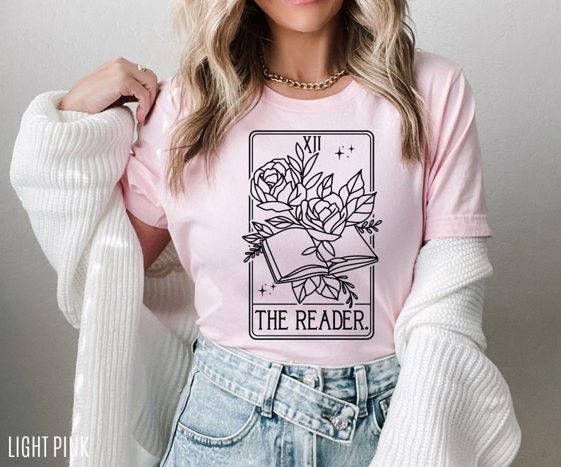 May include: Light pink t-shirt featuring a black and white tarot card design. The design includes an open book, roses, and the text "THE READER." The shirt is made of soft material.