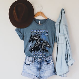 K&ouml;nnte beinhalten: Ein blaues T-Shirt mit einem schwarzen Drachen-Motiv und dem Text "FOURTH WING" in blauen und wei&szlig;en Buchstaben.