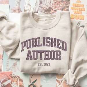 Puede incluir: Una sudadera beige con el texto "PUBLISHED AUTHOR EST. 2023" en morado.