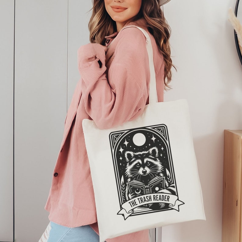 May include: Cream-colored tote bag featuring a black and white illustration of a raccoon reading a book under a night sky with stars and a moon. The text "THE TRASH READER" is below the raccoon.