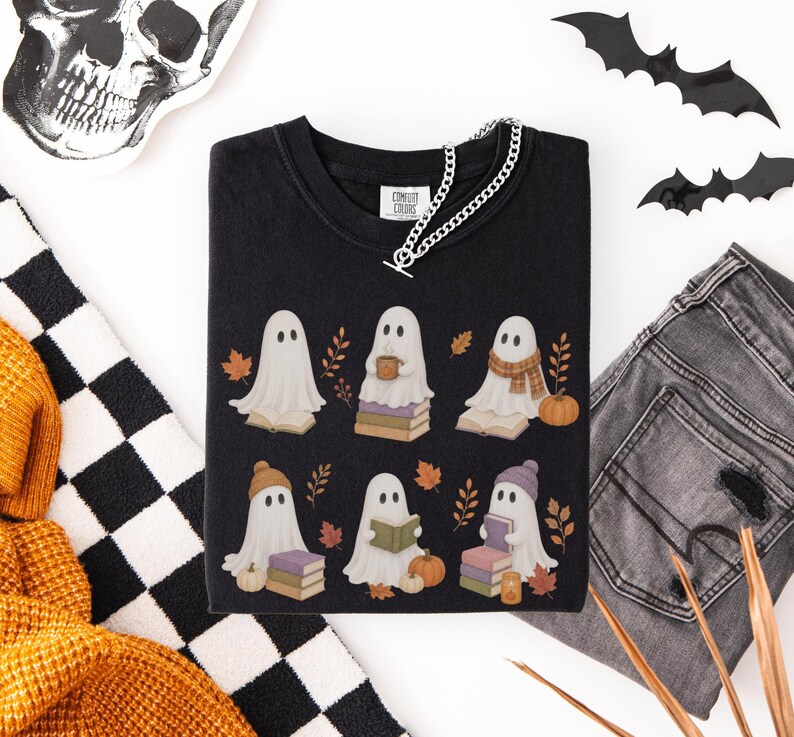 Black shirt featuring six cute bookish ghosts reading books with pumpkins, stacked books, fall leaves, scarves, hats, and a cozy drink.