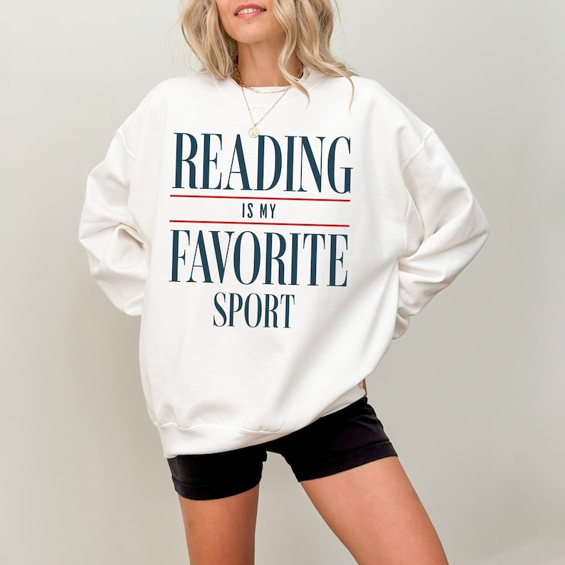 May include: Cream-coloured sweatshirt featuring the navy blue and red text: "READING IS MY FAVORITE SPORT". A comfortable, casual top for anyone who loves reading and books.
