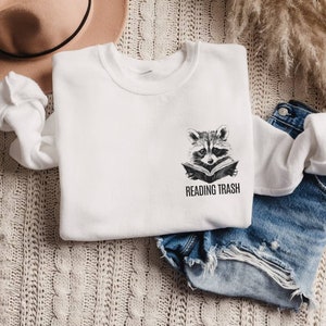May include: White sweatshirt with a raccoon graphic reading a book, with the text "READING TRASH" below. The sweatshirt is folded on a textured, cream-colored surface, with a tan hat and denim shorts nearby.