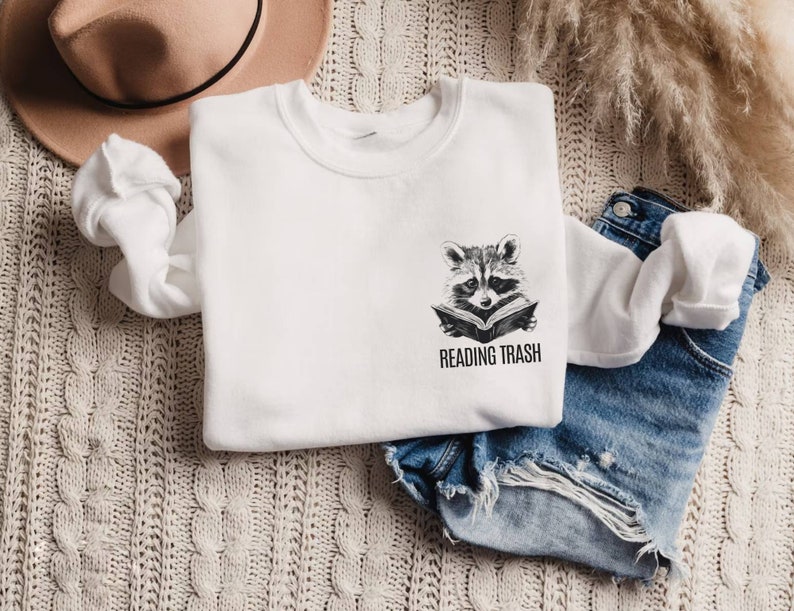 May include: White sweatshirt with a raccoon graphic reading a book, with the text "READING TRASH" below. The sweatshirt is folded on a textured, cream-colored surface, with a tan hat and denim shorts nearby.